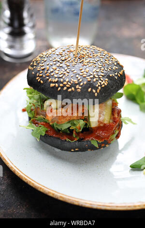Burger in einem schwarzen Bun mit gehacktem Fleisch, Gurken und Ketchup auf einem Teller serviert. Stockfoto