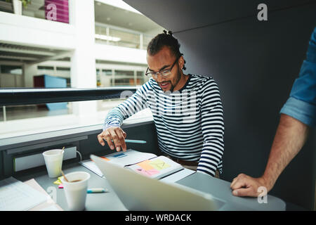 Junge afrikanische amerikanische Geschäftsmann über Noten während der Zusammenarbeit mit Kolleginnen und Kollegen in einem Büro pod konzentriert Stockfoto