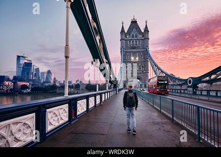 Junger Mann mit Rucksack zu Fuß auf der Tower Bridge gegen Stadtbild mit skyscrapes auf Bunte sunrise. London, Vereinigtes Königreich Stockfoto