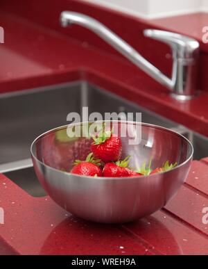 Erdbeeren in einem Edelstahl Schüssel neben einem Spülbecken in leuchtend roten Arbeitsplatte gesetzt. Stockfoto