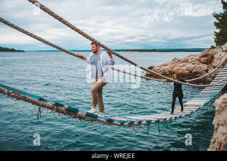 Paar Frau mit Mann zu Fuß durch wackelige Hängebrücke überqueren Sea Bay Stockfoto