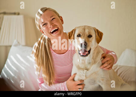 Glückliche junge Frau lacht wie Sie umarmt ihren Hund in Ihrem lichtdurchfluteten Schlafzimmer. Stockfoto