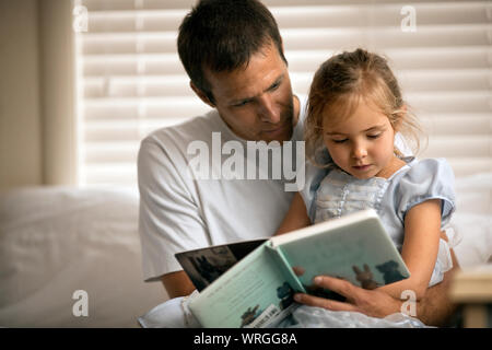 Vater liest sein kleines Mädchen eine Geschichte, wie sie auf seinem Schoß sitzt. Stockfoto