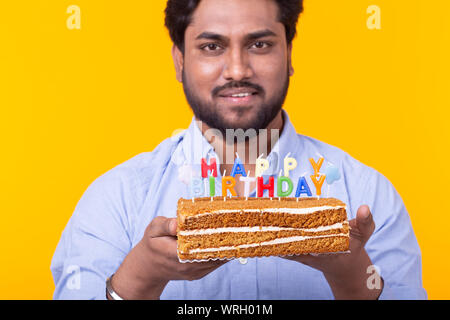 Positive junge Mann hält ein Happy birthday cake und zwei brennende bengalische Lichter auf gelbem Hintergrund posiert. Werbeflächen Stockfoto