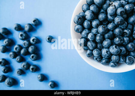 Nahaufnahme einer Platte mit Blaubeeren und in der Nähe von Beeren sind auf blauem Hintergrund verstreut. Quelle der Vitamine und Ernährung Lebensmittel. Stockfoto