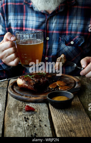 Nahaufnahme der brutalen Grauhaarige erwachsenen Mann mit einem Bart Senf Steak isst und trinkt Bier, das Konzept eines Urlaubs, Festival, Oktoberfest oder St. Patrick's Da Stockfoto