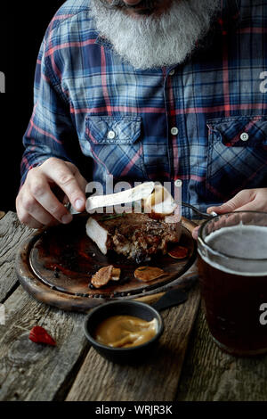 Nahaufnahme der brutalen Grauhaarige erwachsenen Mann mit einem Bart Senf Steak isst und trinkt Bier, das Konzept eines Urlaubs, Festival, Oktoberfest oder St. Patrick's Da Stockfoto