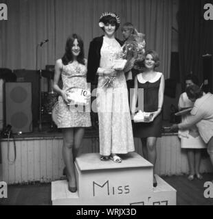 1967, historische, die drei Finalisten in einem lokalen Dorf 'Miss Teenager "Schönheitswettbewerb, stehend auf einem Podium, mit der gewinnenden Mädchen in eine Blume gemusterten Hosenanzug, eine Krone trägt und halten einen Blumenstrauß, England, UK. Stockfoto