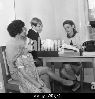 1970 s, historische, eine Mutter mit ihren beiden kleinen Kindern sitzen mit einem weiblichen Sekretär oder Rezeption in einem Büro, England, UK. Stockfoto