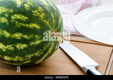 Reif gestreifte Wassermelone mit getrockneten Schwanz und einem langen Messer auf einem braunen Holz Schneidebrett. Cut Wassermelone. Beeren und Früchte, vegetarisches Essen. Stockfoto