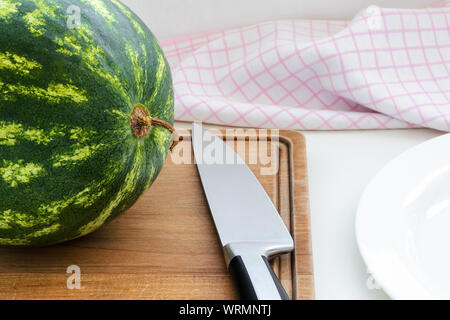 Reif grün gestreiften Wassermelone mit getrockneten Schwanz und einem langen Messer auf einem braunen Holz Schneidebrett. Schneiden Sie die Wassermelone. Beeren und Früchte, veggie. Stockfoto