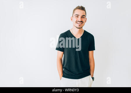 Glückliche junge Kerl in schwarzen T-Shirt auf weißem Hintergrund isoliert mit den Händen in den Taschen posieren. Stockfoto