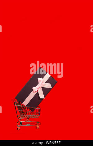 Urlaub Geschenk, Schwarzer Freitag Konzept mit Geschenkbox und rosa Schleife in einem Metall Einkaufswagen, Korb auf rote Farbe Hintergrund. Stockfoto