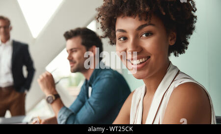 Gefühl glücklich. Junge afro-amerikanische Frau schaut sich Kamera und lächelnd, während mit Kollegen zusammen arbeiten im Büro. Teamarbeit. Konferenz Stockfoto