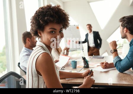 Junge afro-amerikanische Frau schaut sich Kamera und lächelnd, während Ihr Chef in der Nähe von Whiteboard und etwas diskutieren mit Team. Teamarbeit. Konferenz Stockfoto