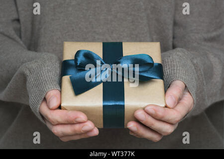 Weibliche Hände im Wollenen beige Pullover, beige Kiste in Handwerk Papier mit blauer Schleife umwickelt. Geschenkset für Mann oder Vater Tag. Close Up. Stockfoto