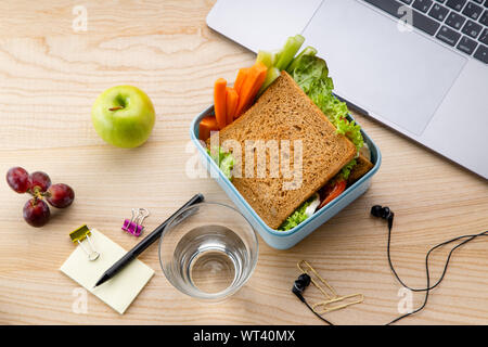 Overhead Bild der gesunde Sandwich in Kunststoffbehälter mit einige Früchte auf hölzernen Tisch neben geöffnet Laptop. Täglich im Büro oder in der Schule Mittagessen Konzept Stockfoto
