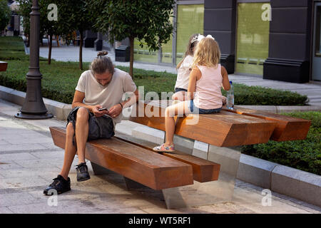 Der Mensch ist auf der Bank sitzen, sein Smartphone aufpaßt, während zwei kleine Mädchen neben ihm spielen Stockfoto