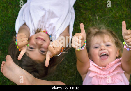 Zwei kleine Mädchen auf dem Gras, lachen und zeigen Daumen hoch Stockfoto