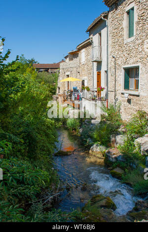 Ein kleiner Fluss, vorbei an den hinteren Türen eines aus Stein gebaute Reihenhäuser in einem französischen Dorf in der Region Drome Stockfoto