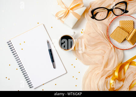 Weibliche Arbeitsbereich mit Notizbuch aus Papier, Stift, Kaffeetasse, Schal, goldenen Accessoires, Band, Geschenk, Waffeln, Gläser auf weißem Hintergrund. Flach, für Frauen Stockfoto