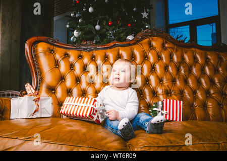 Glückliche kleine lächelnde Junge mit Weihnachten Geschenkverpackung. Schönes kleines Baby, sitzt auf asofa mit Geschenk. Kind sitzen durch einen Stapel Weihnachtsgeschenke Stockfoto