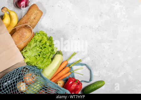 Frischen landwirtschaftlichen Gemüse in Netztasche und Brot, Baguettes und Bananen in Papiertüte auf grauem Beton Hintergrund. Ansicht von oben, horizontale Ausrichtung, Kopieren spac Stockfoto