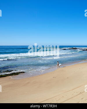 Blick auf den Strand mit einer Person in der Ferne wandern entlang der Gewässer Kante wie die Flut kommt auf einem sonnigen, wolkenlosen Herbst Tag in Australien Stockfoto