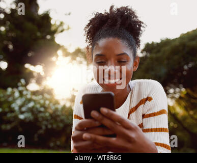 Portrait einer afrikanischen amerikanischen Jungen Frau mit Handy im Park an einem sonnigen Tag lächelnd - sehr glücklich Frau auf Handys Stockfoto