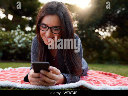 Glückliche junge Frau das Tragen von Brillen liegen auf Decke über Gras im Park mit Handy Stockfoto