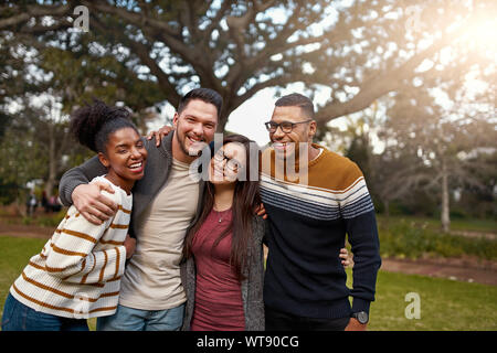 Lächelnd Gruppe von Freunden zusammen lachen, während mit Arme um einander in einem Park stehen - glückliche Gruppe von Menschen Stockfoto