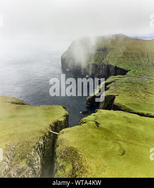 Graue Wolken über den felsigen Klippen mit grünem Moos in der Nähe von ruhigen Meer an trüben Tag auf den Färöer Inseln Stockfoto