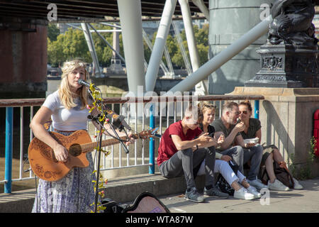 Busker Sänger mit Gitarre auf der Southbank, London mit einer Familie von Touristen sitzen gegen ein Geländer der Themse, etwas zu Essen. Stockfoto