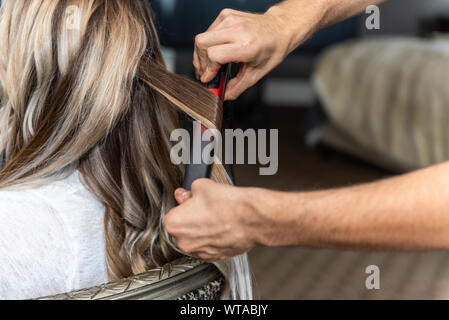 Die langen Haare der Frau in Stuhl mit geschickten Händen mit einem Lockenstab gestaltet wird, sitzt. Stockfoto