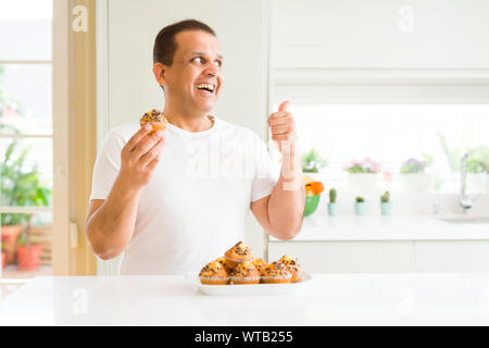 Mittleres Alter Mann essen Chocolate Chip Muffins zu Hause zeigen und zeigt mit dem Daumen bis zu der Seite mit glücklichen Gesicht lächelnd Stockfoto
