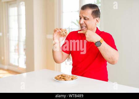 Mittleres Alter Mann essen Chocolate Chips cookies zu Hause sehr zufrieden zeigte mit Hand und Finger Stockfoto
