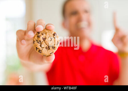 Mittleres Alter Mann essen Chocolate Chips cookies zu Hause mit einer Idee oder Frage Zeigefinger mit glücklichen Gesicht überrascht, Nummer eins Stockfoto