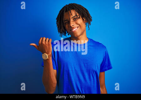 Afro-amerikanische Mann mit Dreadlocks tragende t-shirt stehend über isolierte blauen Hintergrund lächelnd mit glücklichen Gesicht und mit Verweis auf die Seite Stockfoto