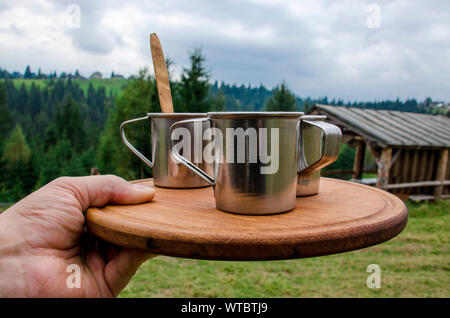Der Mann hält ein Fach mit drei Tassen Kaffee auf einem Holzbrett auf einem Hintergrund von Wald und Berge Stockfoto