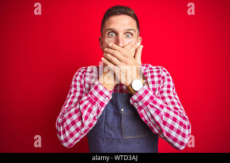 Jungen gutaussehenden Mann, der Ladenbesitzer Schürze einheitlich über den roten Hintergrund isoliert schockiert über den Mund mit Händen für Fehler. Geheime Konzept. Stockfoto