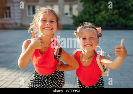 Glückliche Schwestern Mädchen, Rucksäcke und Daumen hoch. Kinder Schüler Spaß im Freien Schule. Bildung. Zurück zur Schule Stockfoto