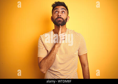 Junge Inder tragende t-shirt stehend über isolierte gelben Hintergrund Denken konzentriert über Zweifel mit dem Finger auf das Kinn und sah wonderi Stockfoto