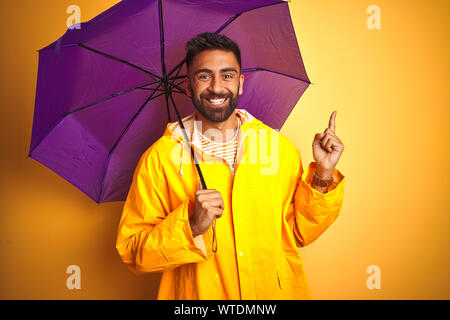 Junge indische Mann mit Regenmantel und lila Schirm über isolierte gelben Hintergrund sehr zufrieden zeigt mit Finger- und an der Seite Stockfoto