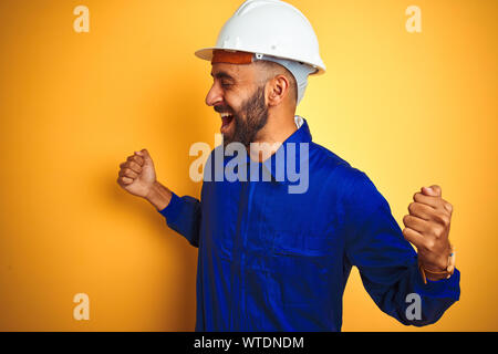 Hübscher indischer Arbeitnehmer Mann einheitliche und Helm tragen über isolierte gelben Hintergrund sehr glücklich und aufgeregt, Sieger Geste mit erhobenen Armen, sm Stockfoto