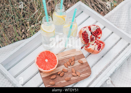 Picknick Set mit Grapefruit, Granatapfel, Mandel- Muttern und Limonade in Glasflaschen auf weißem Holz- Fach. Stockfoto