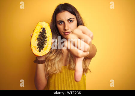 Junge schöne Frau mit Papaya über gelb isoliert Hintergrund mit Verärgerten Gesicht, negatives Vorzeichen zeigen Abneigung mit Daumen nach unten, Ablehnung von quantitativen Simulatio Stockfoto