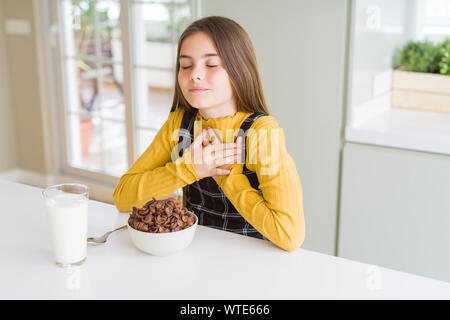 Schöne junge Mädchen Junge Schokolade essen Getreide und Glas Milch zum Frühstück lächelnd mit der Hand auf der Brust mit geschlossenen Augen und dankbare Geste auf Stockfoto