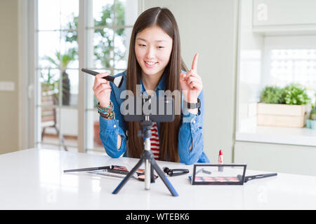 Schöne asiatische Einflussnehmer Frau Aufnahme Make-up Tutorial mit Smartphone Kamera mit einer Idee oder Frage Zeigefinger mit glücklichen Gesicht überrascht Stockfoto