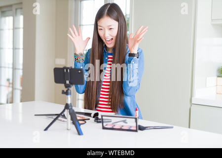 Schöne asiatische Einflussnehmer Frau Aufnahme Make-up Tutorial über Smartphone-kamera verrückt und crazy für den Erfolg mit erhobenen Armen und geschlossenen Feiern Stockfoto