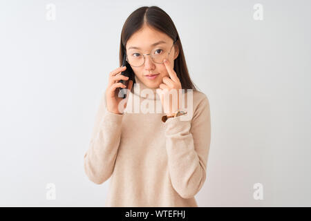 Junge chinesische Frau sprechen über das Smartphone über isoliert weißer Hintergrund, der auf das Auge beobachtet sie Geste, verdächtige Ausdruck Stockfoto